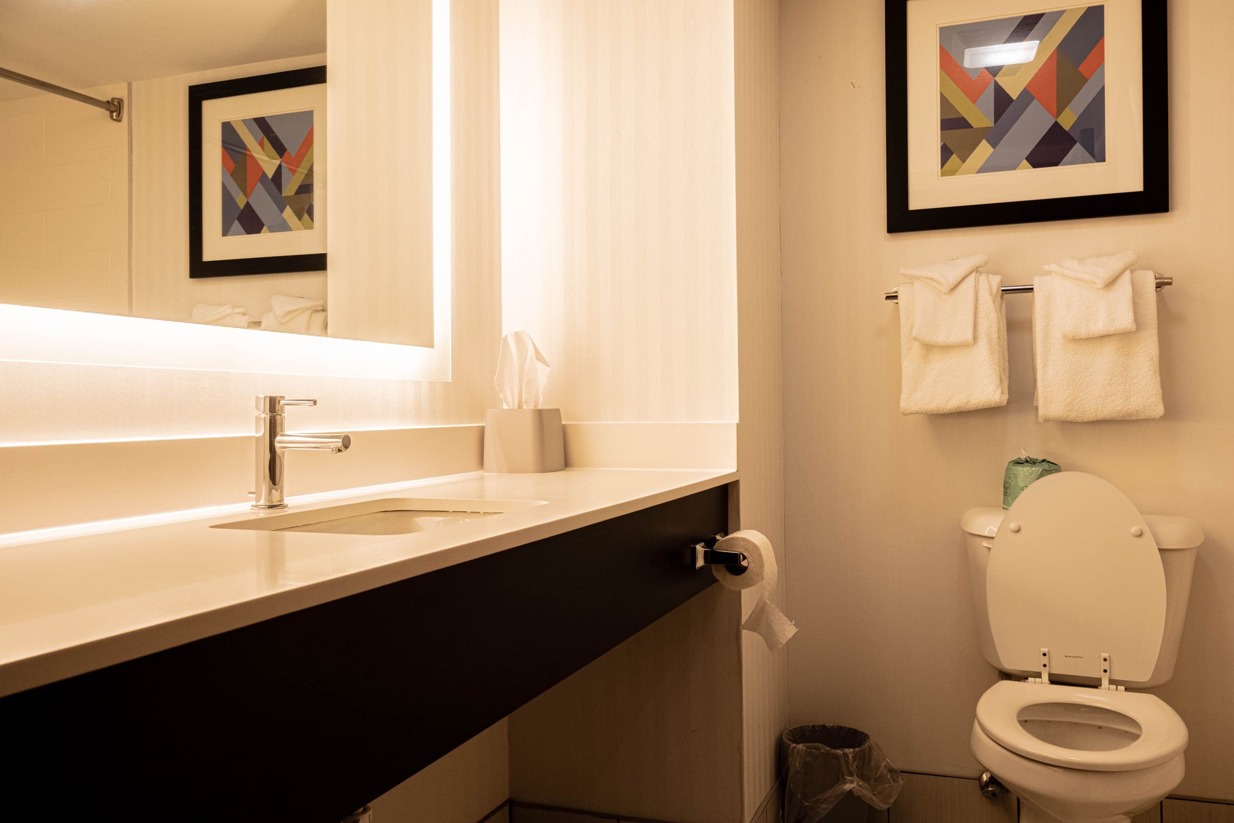 Hotel bathroom with a backlit mirror, quartz vanity, folded towels, and framed geometric artwork